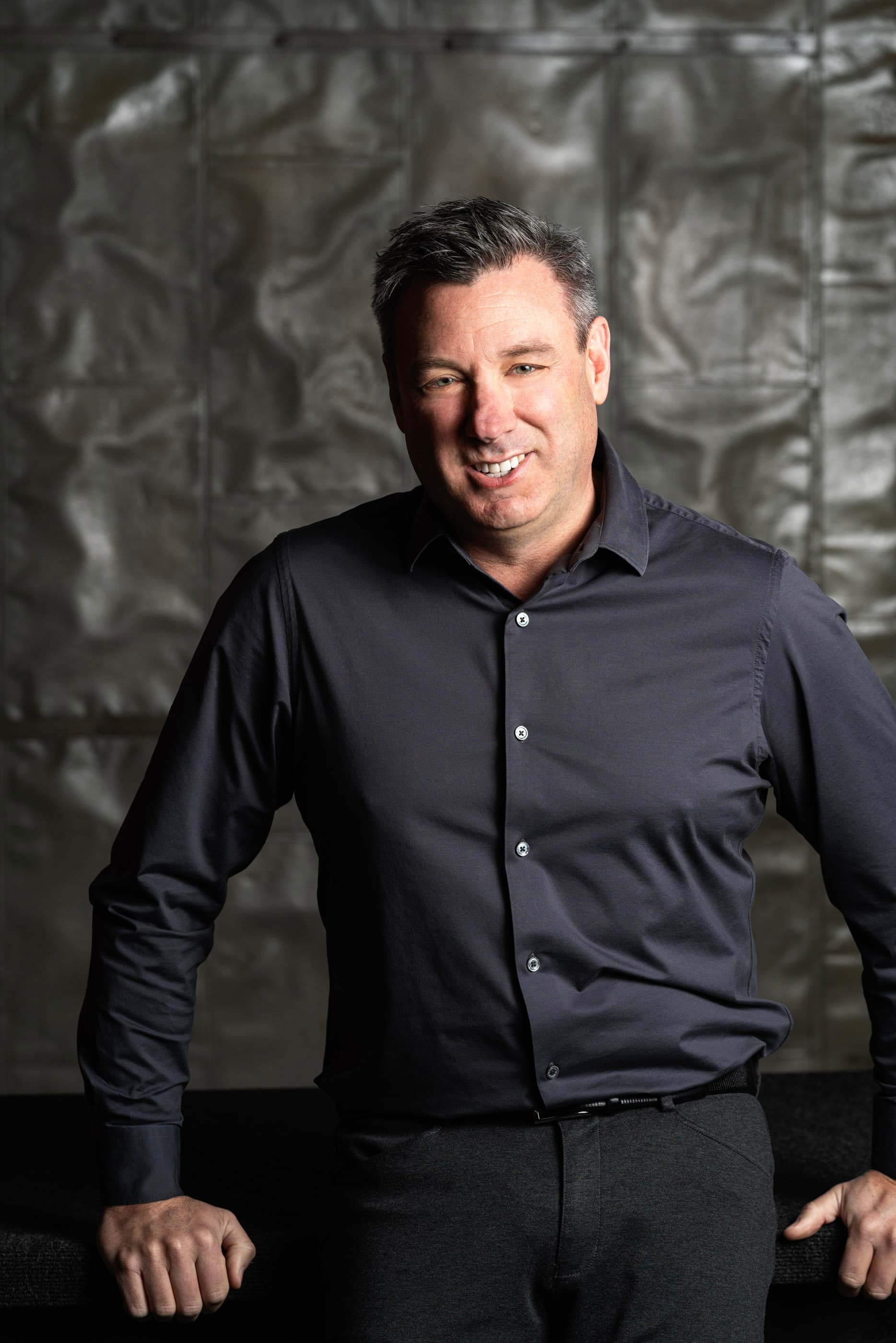 Smiling man in a dark gray button-down shirt posing against a textured metallic wall.