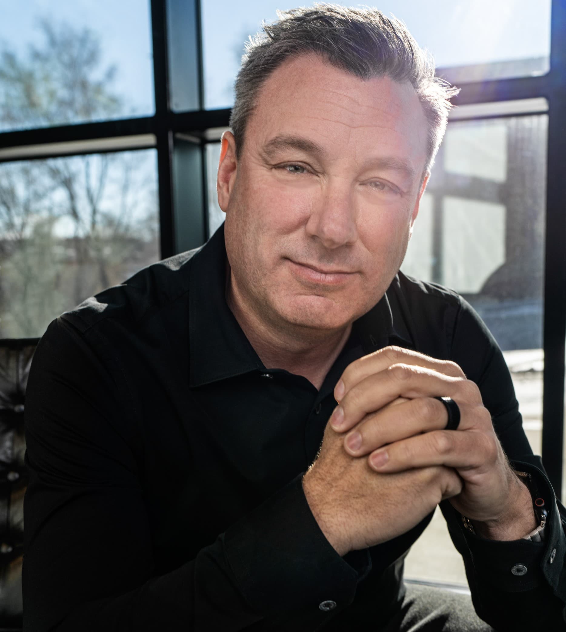 Middle-aged man in a black shirt with hands clasped, sitting in front of a window.